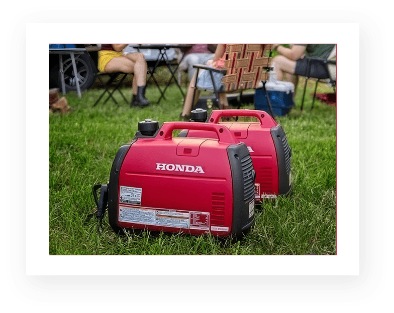 Two red generators sitting in the grass near a group of people.